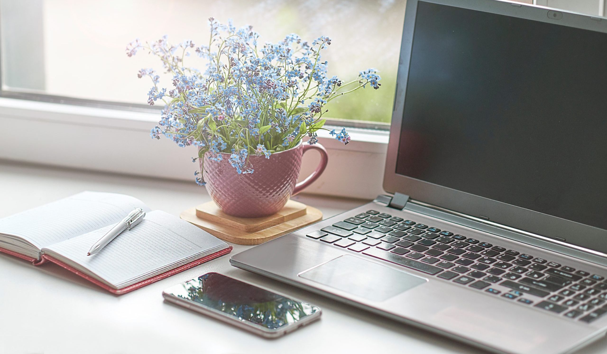 An open notebook, cellphone, laptop and flowers on a table.