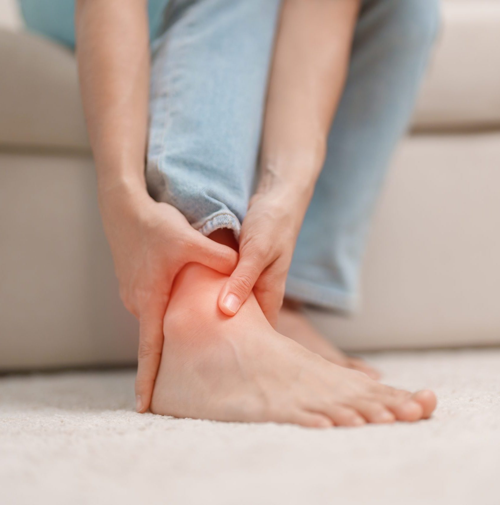 A woman sitting on her couch feeling her right ankle highlighted in red to indicate pain.
