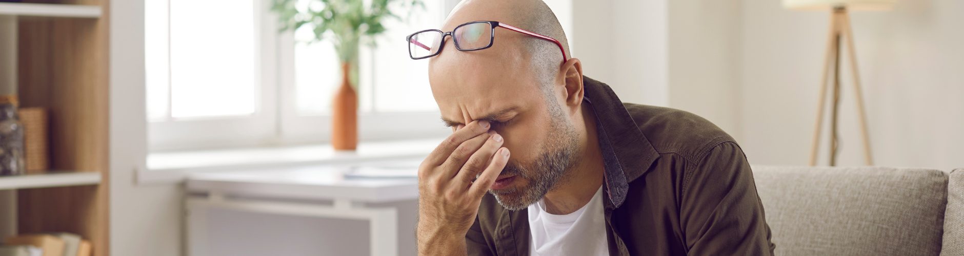 Man sitting on a couch with glasses resting on his head, rubbing his eyes and appearing fatigued or stressed.