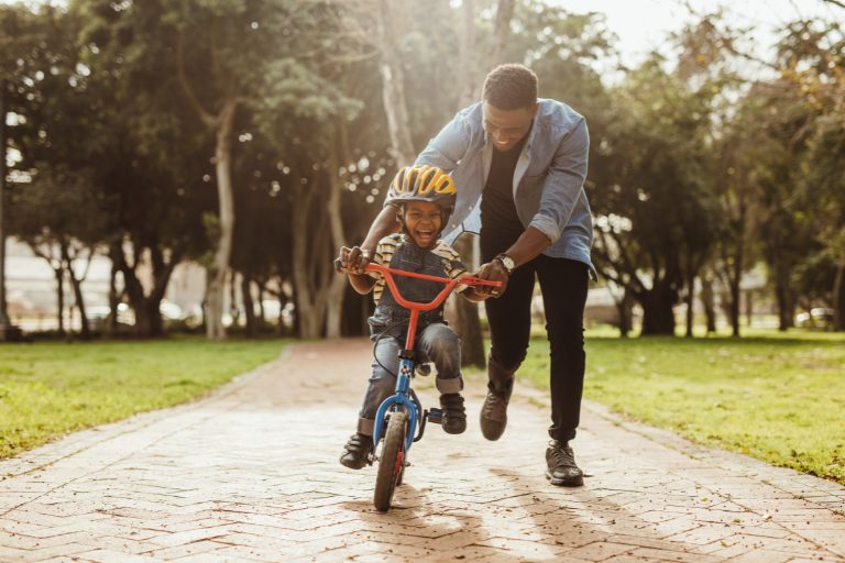 Adult helping a child learn to ride a bicycle on a park path while the child wears a helmet